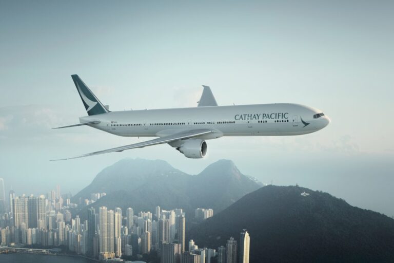 Cathay Pacific wide-body airliner flying above a coastal city with mountains in the background, under a clear sky.