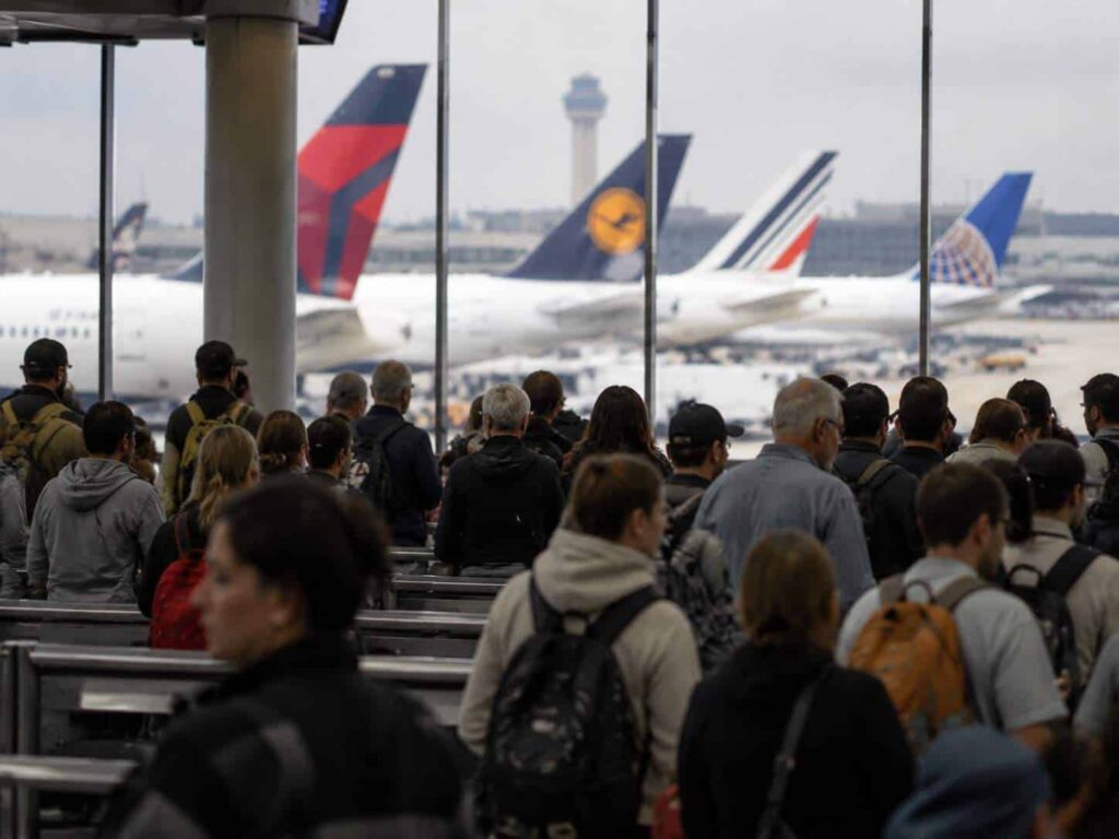 Crowd of travelers with backpacks waiting in a busy airport terminal, with several jetliners visible through large windows outside.