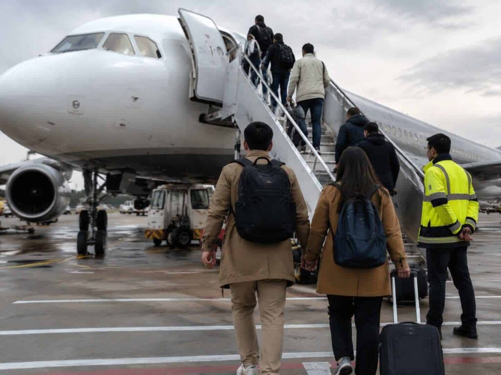 Passengers with rolling luggage walk up the mobile stairs to board a large white airplane on a wet tarmac, with a ground crew member in a high‑vis vest nearby.
