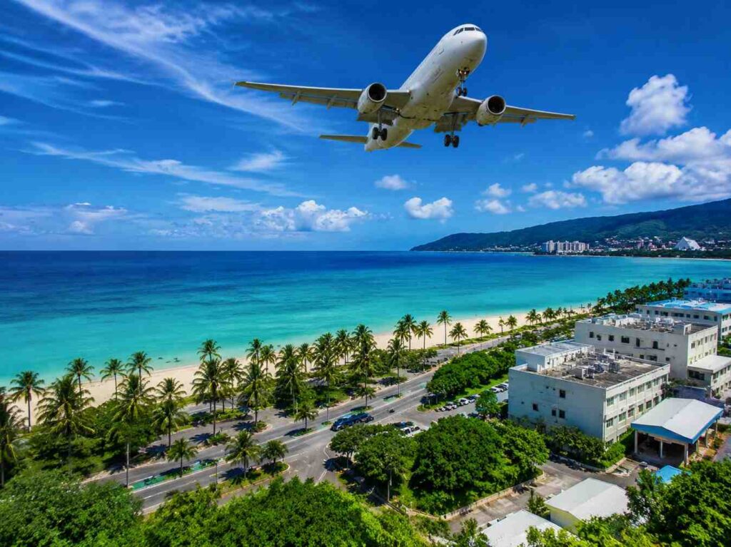 Airplane flying low over a tropical beach with turquoise water, palm trees lining the shore, and white buildings nearby.