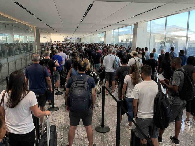 Crowded airport terminal with dozens of travelers in line, many carrying backpacks and rolling luggage behind black stanchions.