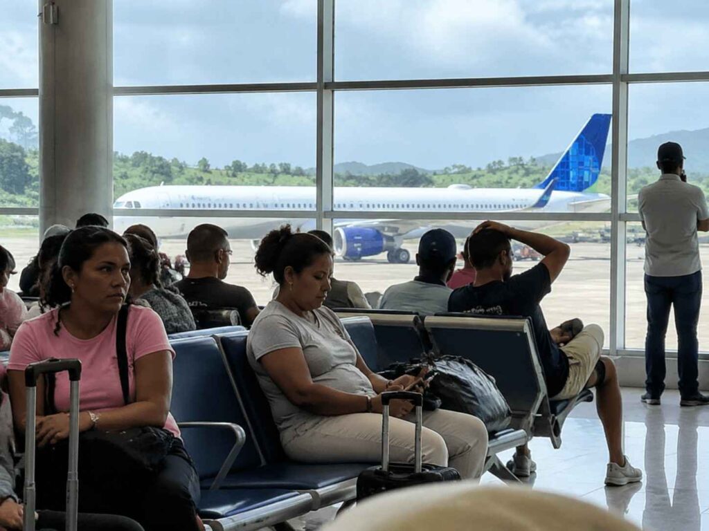 Passengers seated in an airport terminal watching a white airplane with a blue tail on the tarmac outside large windows.