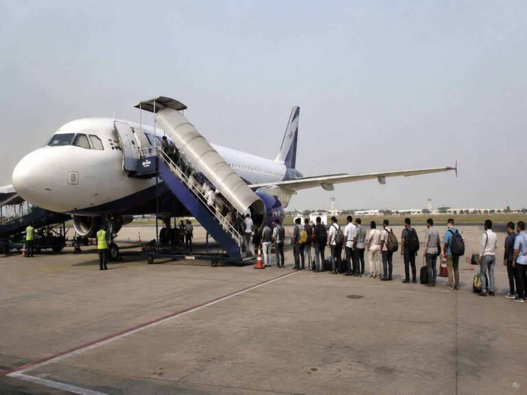 White passenger jet parked on tarmac with mobile boarding stairs attached and passengers lined up to board, carrying backpacks and bags under a clear sky