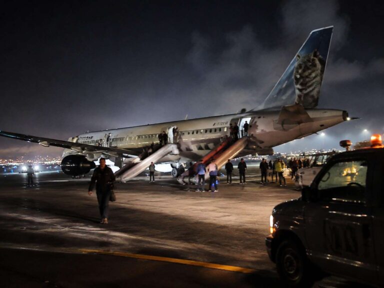 Night-time airport scene: passengers boarding a large jet via mobile stairs, with ground crew and a vehicle nearby and city lights in the distance.
