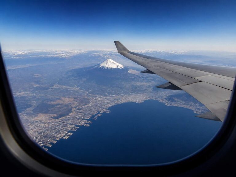 Airplane window view with the wing in the foreground and a snow‑capped Mount Fuji visible beyond the coast below.