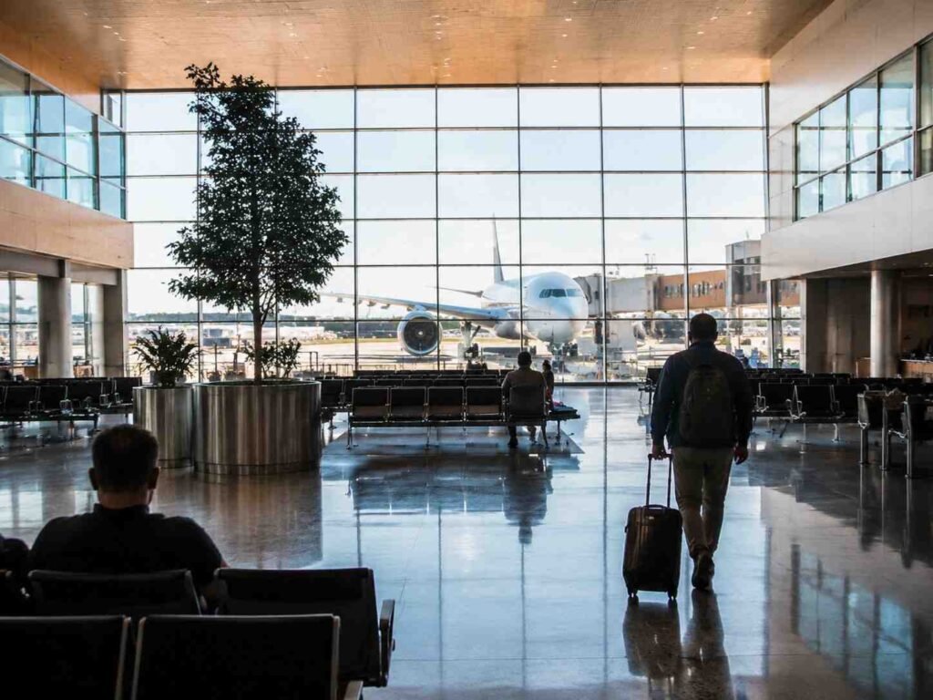 Airport terminal with a large glass wall; a jet at the gate outside and travelers seated inside, a man with a rolling suitcase walking toward the plane.