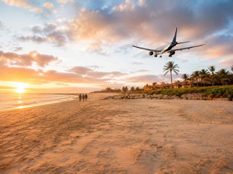 Sunset beach scene with three people walking along the shore as a plane approaches for landing; palm trees and a resort line the coast.