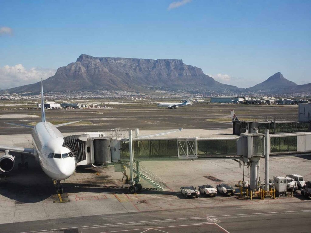 White passenger jet at a gate with jet bridge, parked on the concrete apron as a distant mountain range and blue sky loom.