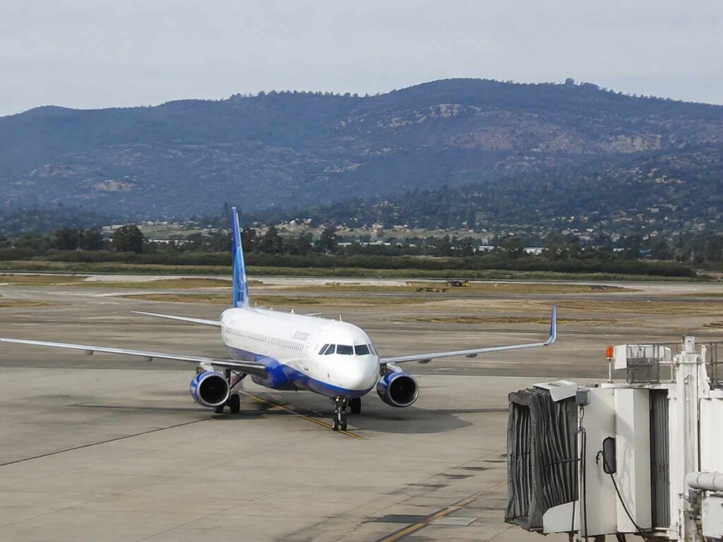 White and blue commercial airplane parked at a gate with a jet bridge connected, on a sunlit tarmac with hills in the background.