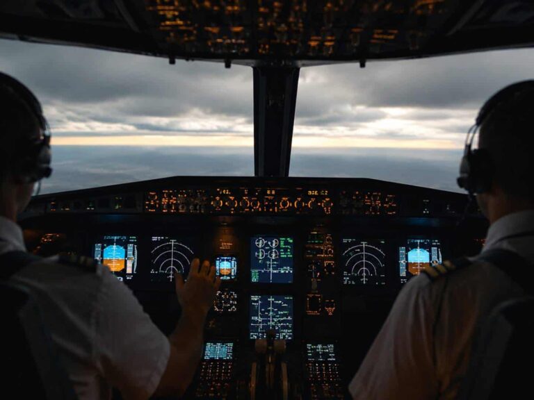 Two pilots in a dim cockpit wearing headsets, monitors glowing with flight instruments and a sunset horizon outside the windshield.