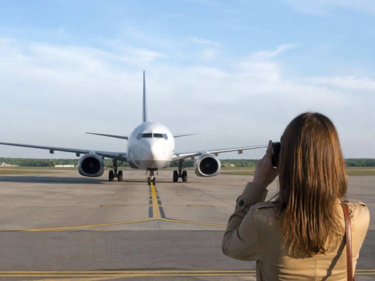 Person with a camera taking a photo of a large white airplane on the runway, seen from behind the photographer's back