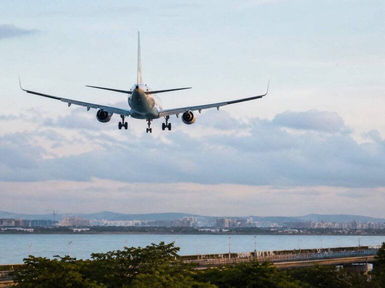 Commercial jet on final approach, wings spread, descending toward a coastal city with water and buildings below