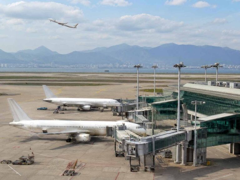 Airplanes parked at a modern terminal with a jet bridge as one plane takes off in the background.