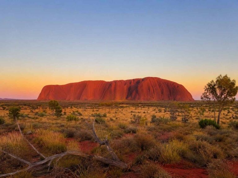 Massive red rock formation Uluru rising from a flat desert plain at sunset, with sparse scrubland in the foreground.