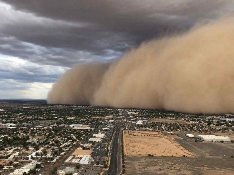 Aerial view of a massive dust storm rolling over a desert city, with a pale orange wall of dust advancing across the streets and rooftops under dark, stormy clouds.