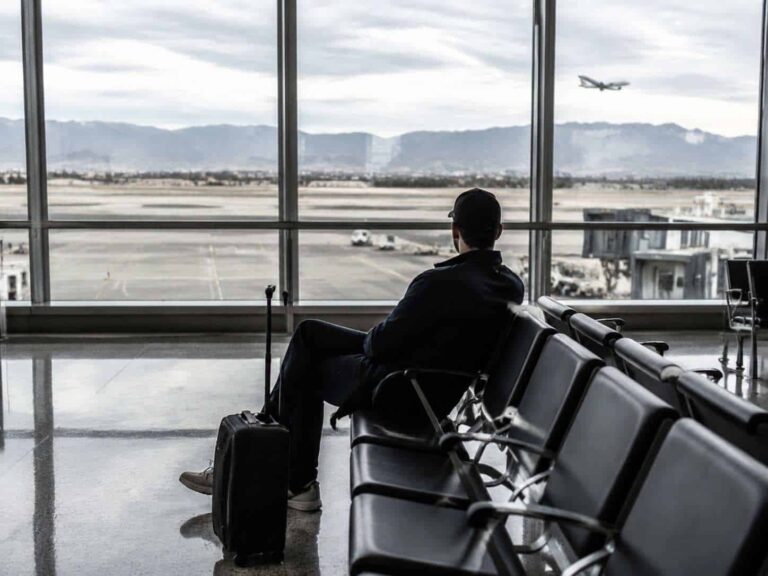Traveler sits with rolling suitcase in an airport terminal, watching a plane take off beyond the tarmac through large windows.