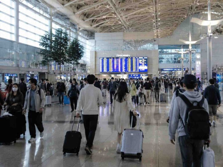 Busy airport terminal with travelers pulling rolling suitcases and a large flight information display in the background.