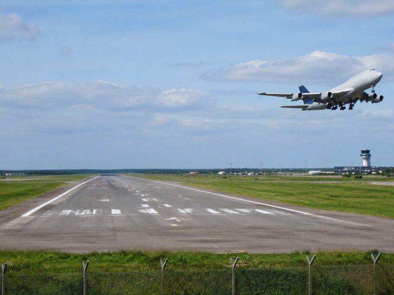 Commercial jet lifting off from a runway at a sunny airport, with a control tower visible in the distance.