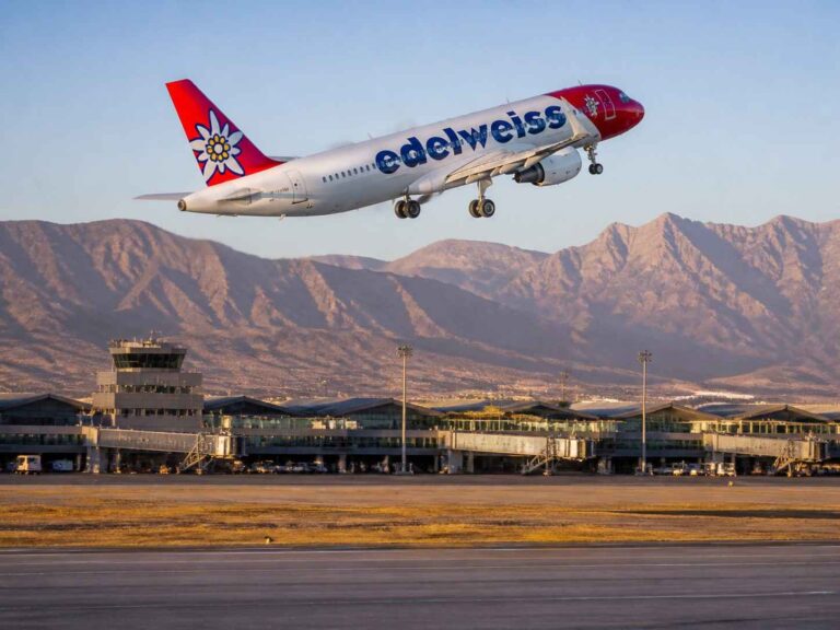 Edelweiss passenger plane takes off from a runway with terminal buildings and desert mountains in the background.
