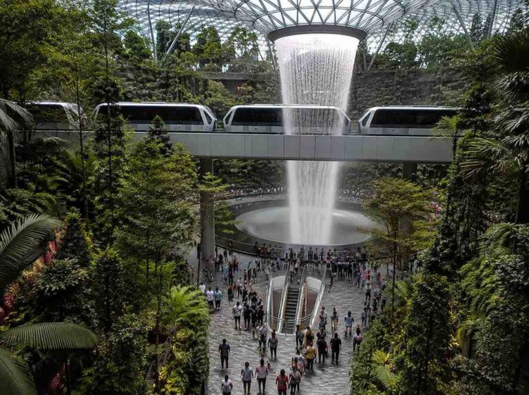 Indoor tropical garden with a tall cascading waterfall, a glass ceiling, and a suspended monorail above a busy visitors' area beneath lush greenery.
