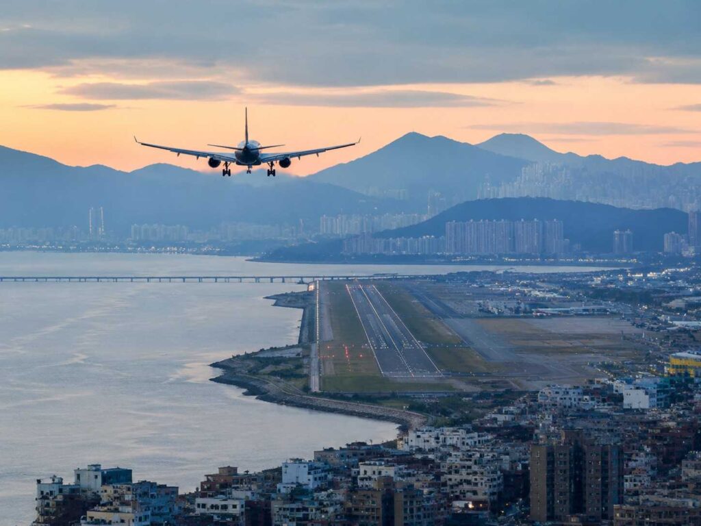 Airplane approaches a coastal runway over water with a city skyline and distant hills at sunset.