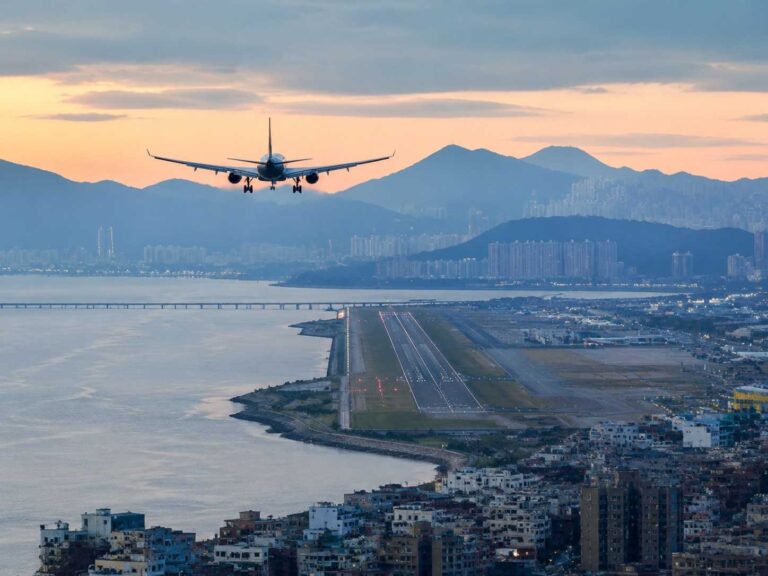 Airplane approaches a coastal runway over water with a city skyline and distant hills at sunset.