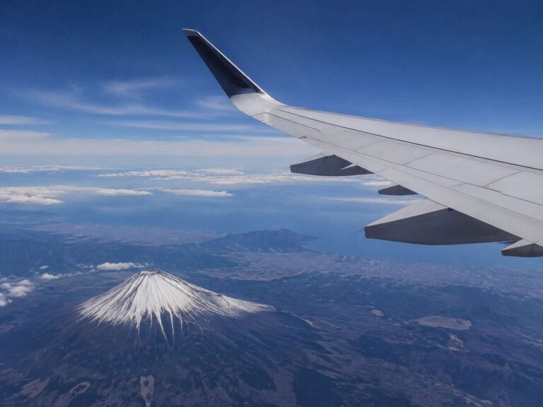 Airplane wing with winglet visible against a blue sky, overlooking a snow-covered volcano and distant coastline below.