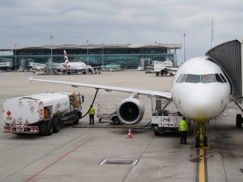 A large white commercial airplane at the gate with a fuel truck connected to its wing and ground crew nearby, terminal in the background.