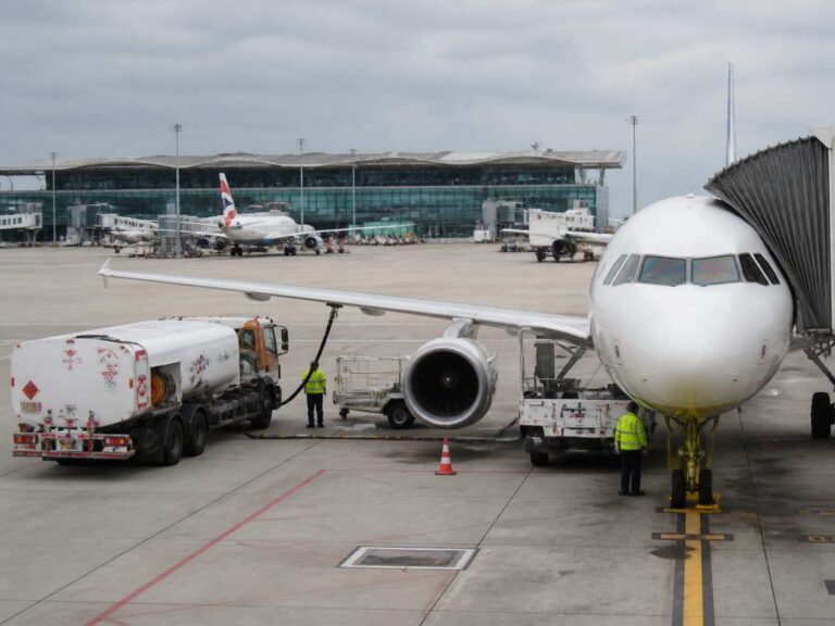 A large white commercial airplane at the gate with a fuel truck connected to its wing and ground crew nearby, terminal in the background.