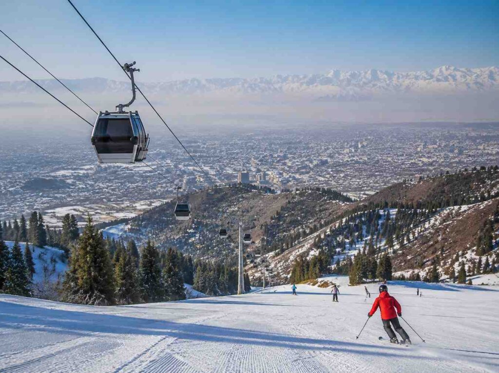 Gondola lift over a snowy ski slope with skiers below and a city in the valley, mountains in the distance.