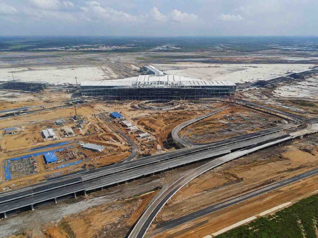 Aerial view of a large construction site with a modern terminal under construction and looping elevated roads nearby, on a sandy landscape water horizon in the distance.