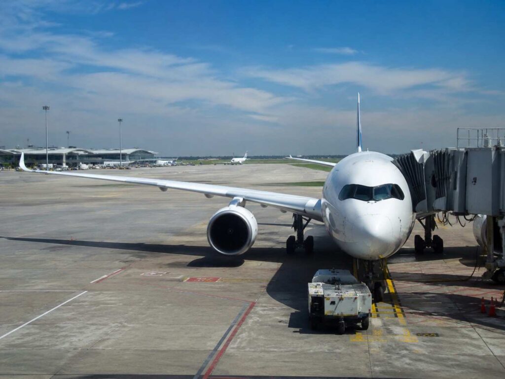 Front view of a white passenger jet parked at a gate with a jet bridge attached on a sunny airport tarmac