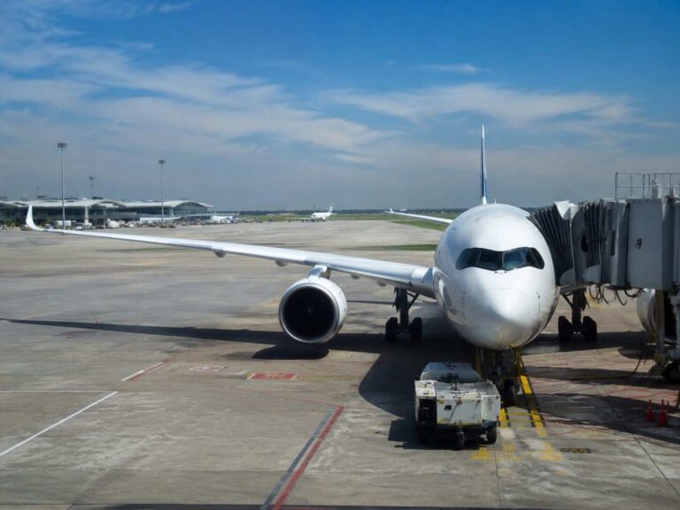 Front view of a white passenger jet parked at a gate with a jet bridge attached on a sunny airport tarmac