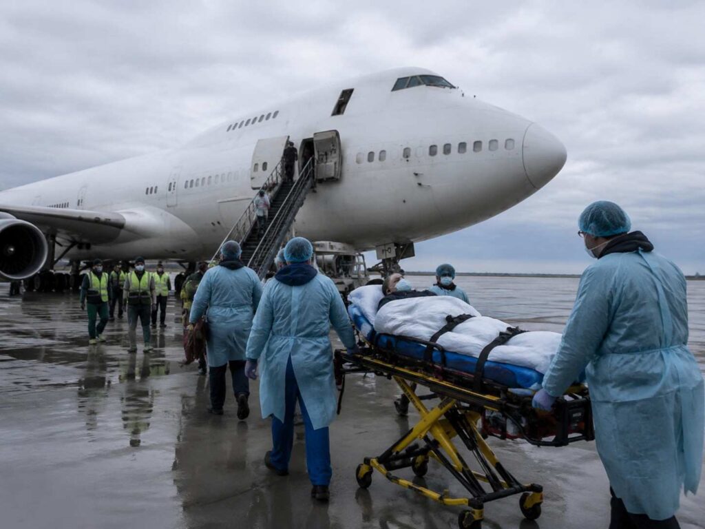 Medical team in blue PPE unloads a patient on a stretcher from a large white plane on a wet tarmac.