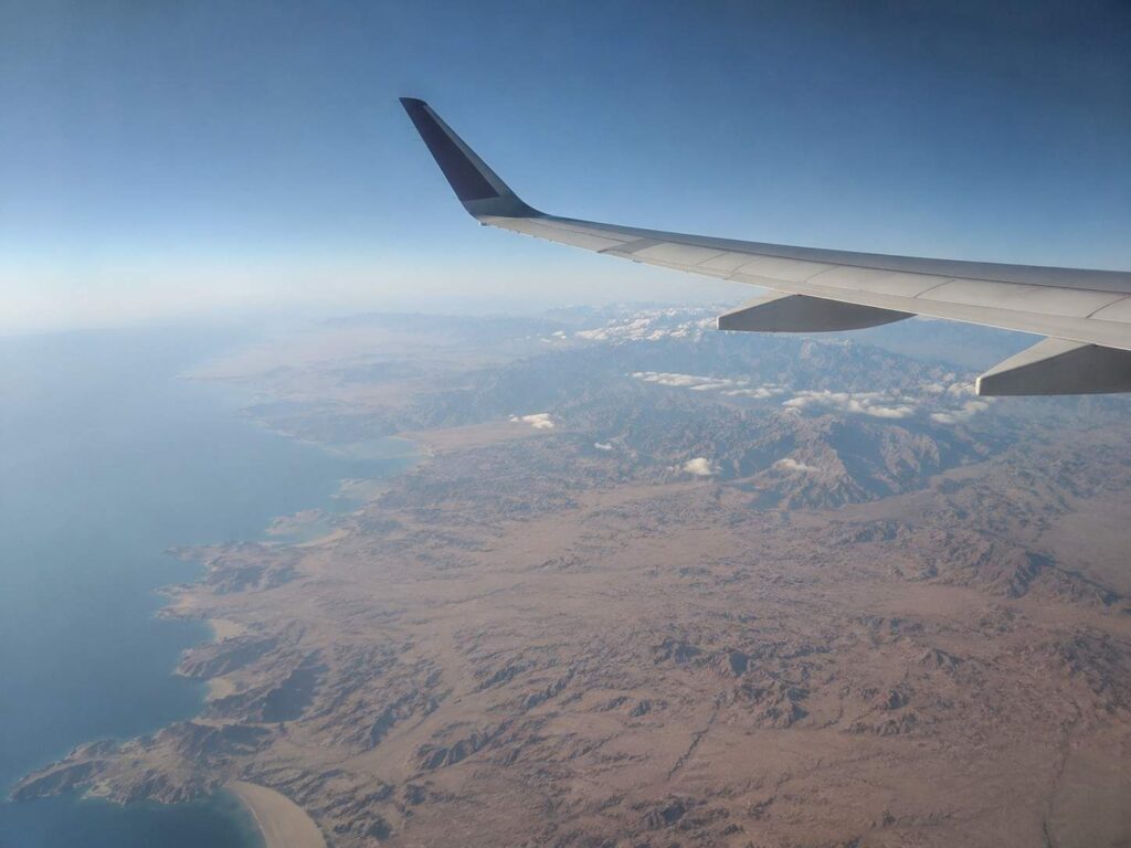 Airplane wing seen from a passenger window above a desert coastline and rugged mountains under a clear blue sky.