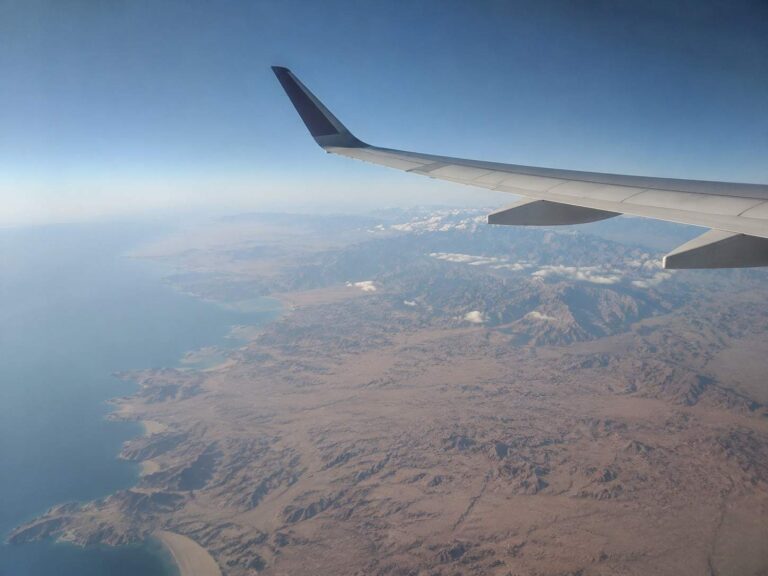 Airplane wing seen from a passenger window above a desert coastline and rugged mountains under a clear blue sky.
