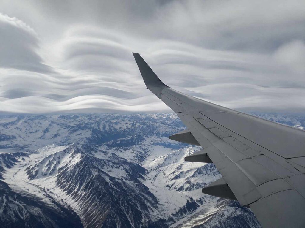 Airplane wing seen from a window over snow-covered mountains with dramatic layered clouds above.