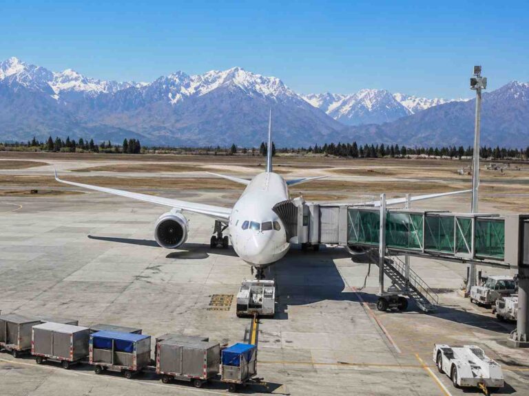 Front view of a white passenger jet at a gate with a jet bridge attached, snow-capped mountains in the distance under a clear blue sky.