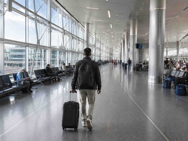 Man in a black jacket pulls a rolling suitcase through a spacious airport terminal with large glass walls and metal support pillars.
