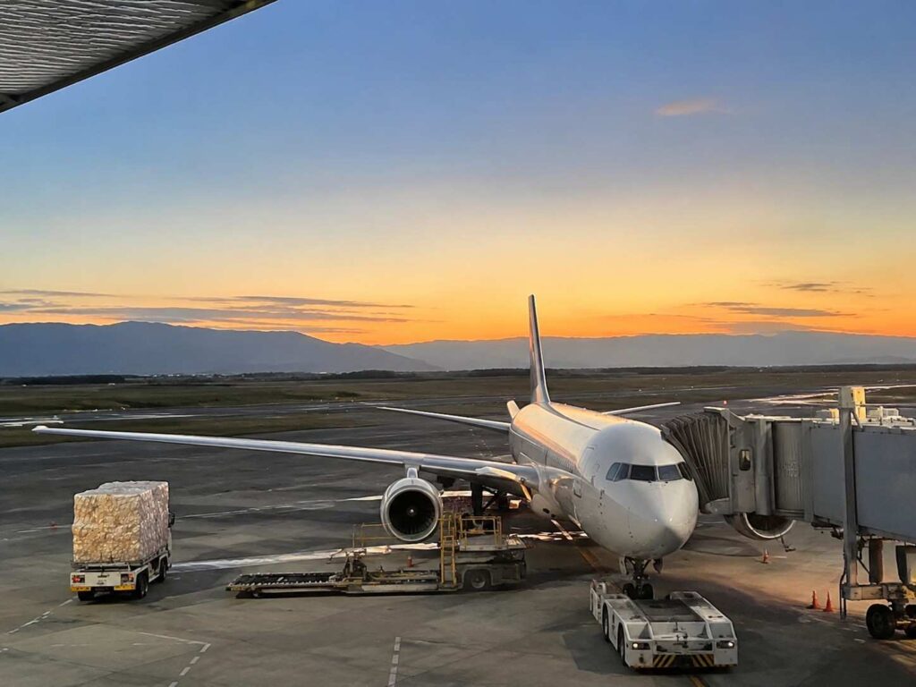 Large white commercial airplane docked at a gate with a jet bridge attached, sunset sky and distant mountains on the horizon, cargo pallet on a truck nearby.