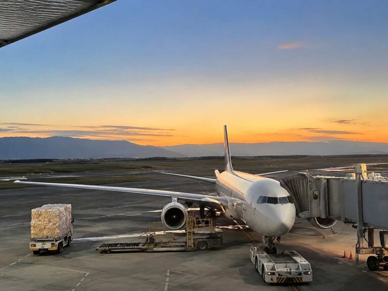 Large white commercial airplane docked at a gate with a jet bridge attached, sunset sky and distant mountains on the horizon, cargo pallet on a truck nearby.