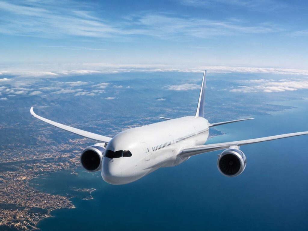 White wide-body airliner flying over a coastline with scattered clouds and blue sky below the wings