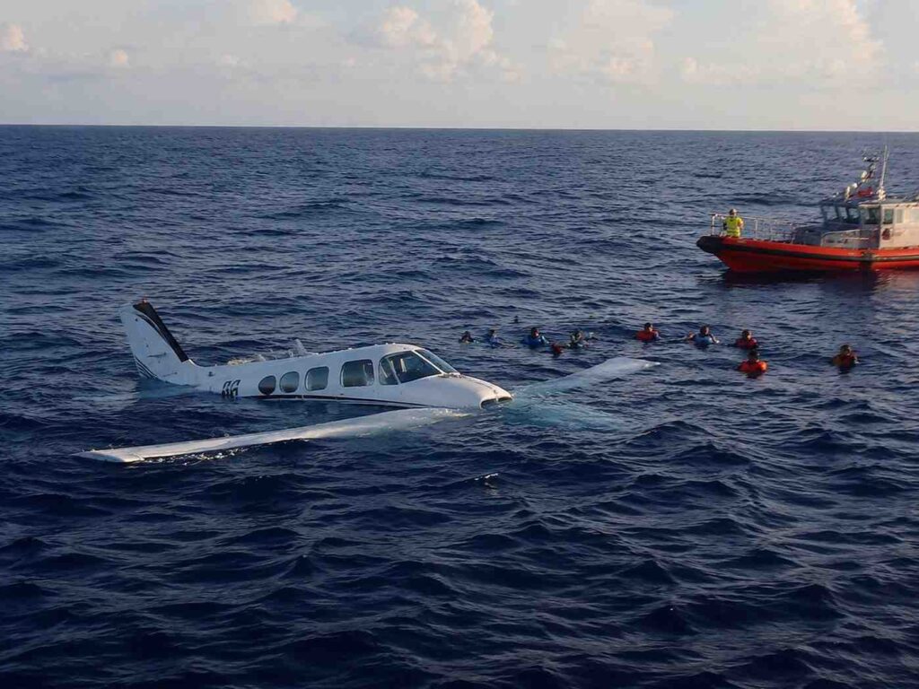 Partially submerged light aircraft wreck in the open sea; swimmers surround the fuselage as a red rescue boat looms on the right.