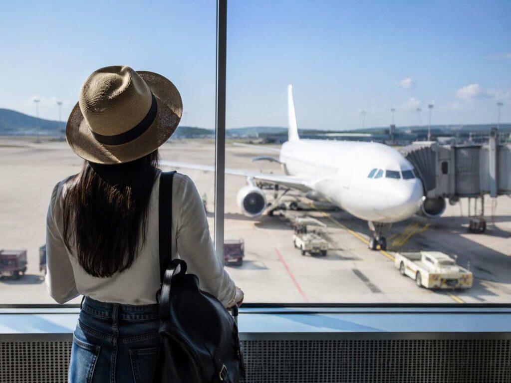 Traveler in a wide-brimmed hat stands at a terminal window, watching a white aircraft dock at the gate outside.