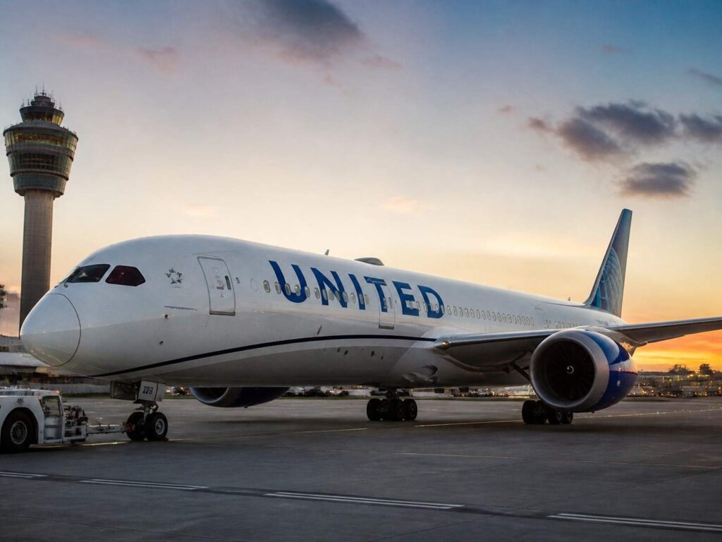 United aircraft on tarmac at sunset, nose gear down, control tower in background and jet engine visible on the right
