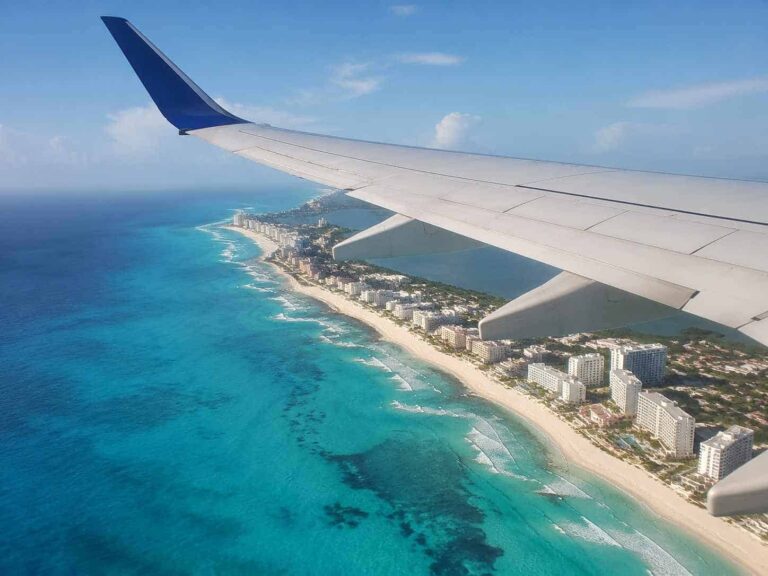 Wing of an airplane over a tropical coastline with turquoise water and white-sand beach below, seen from a window seat.