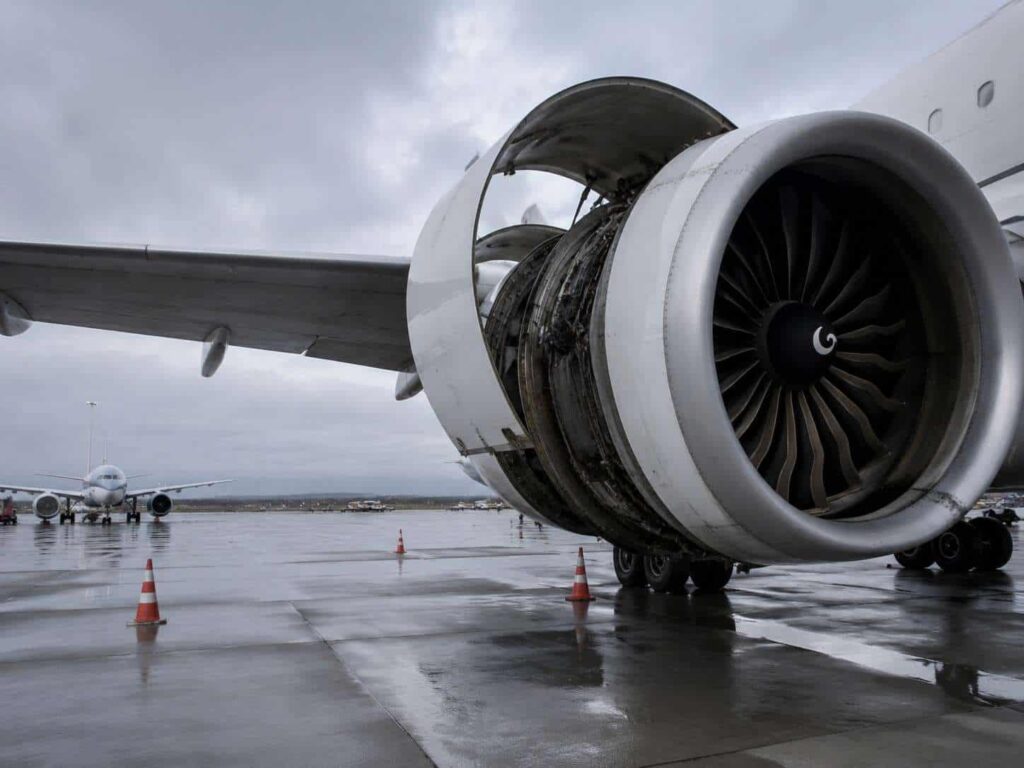 Close-up of a jet engine with its cowling removed and exposed turbine blades on a wet tarmac, safety cones nearby.