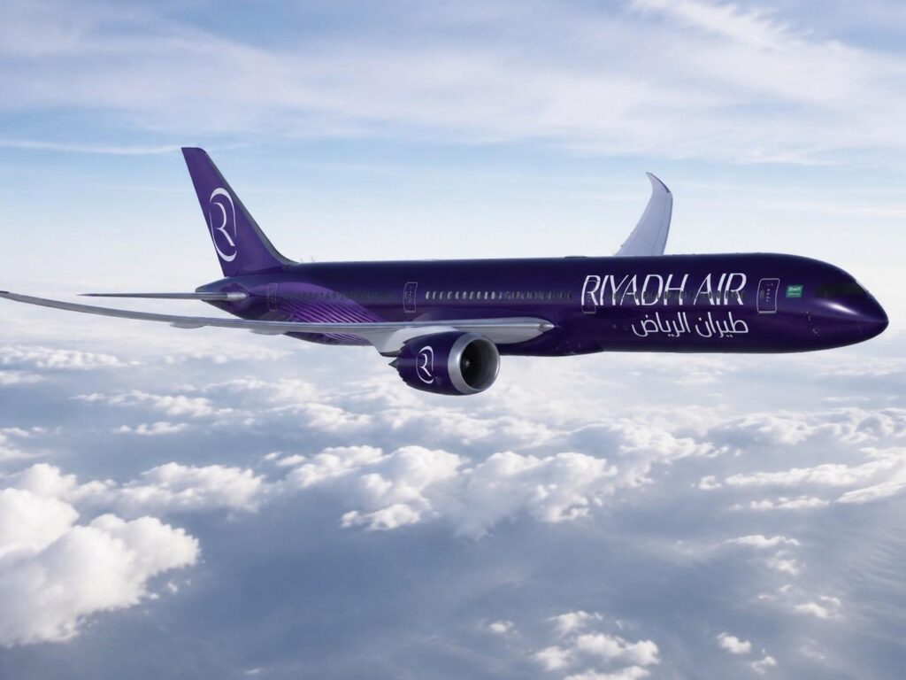 Purple Riyadh Air jet in flight above white clouds, with Arabic and English branding on the fuselage and a white vertical stabilizer in the background.