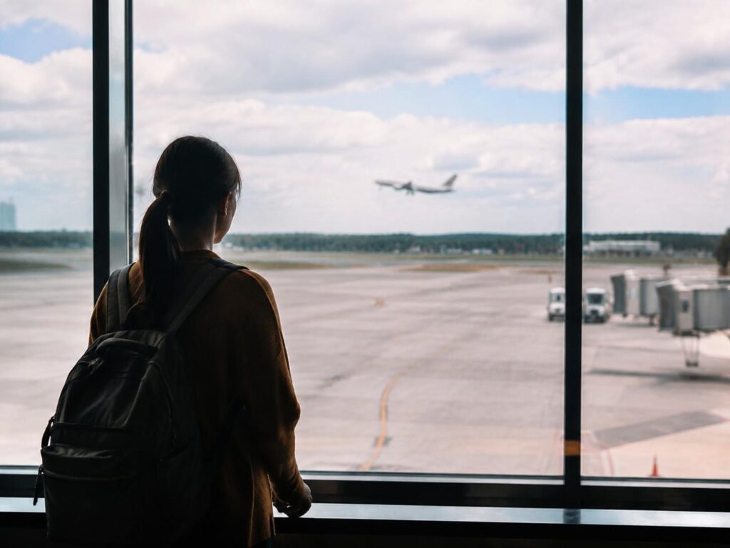 Traveler with a backpack stands at a large window, watching a plane take off on the runway.
