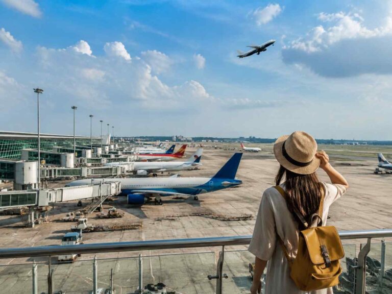 Traveler with a straw hat and tan backpack stands on an airport viewing deck, watching planes at gates below.